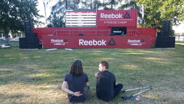 Two people sitting on the grass opposite the Reebok Fitness Camp stage.