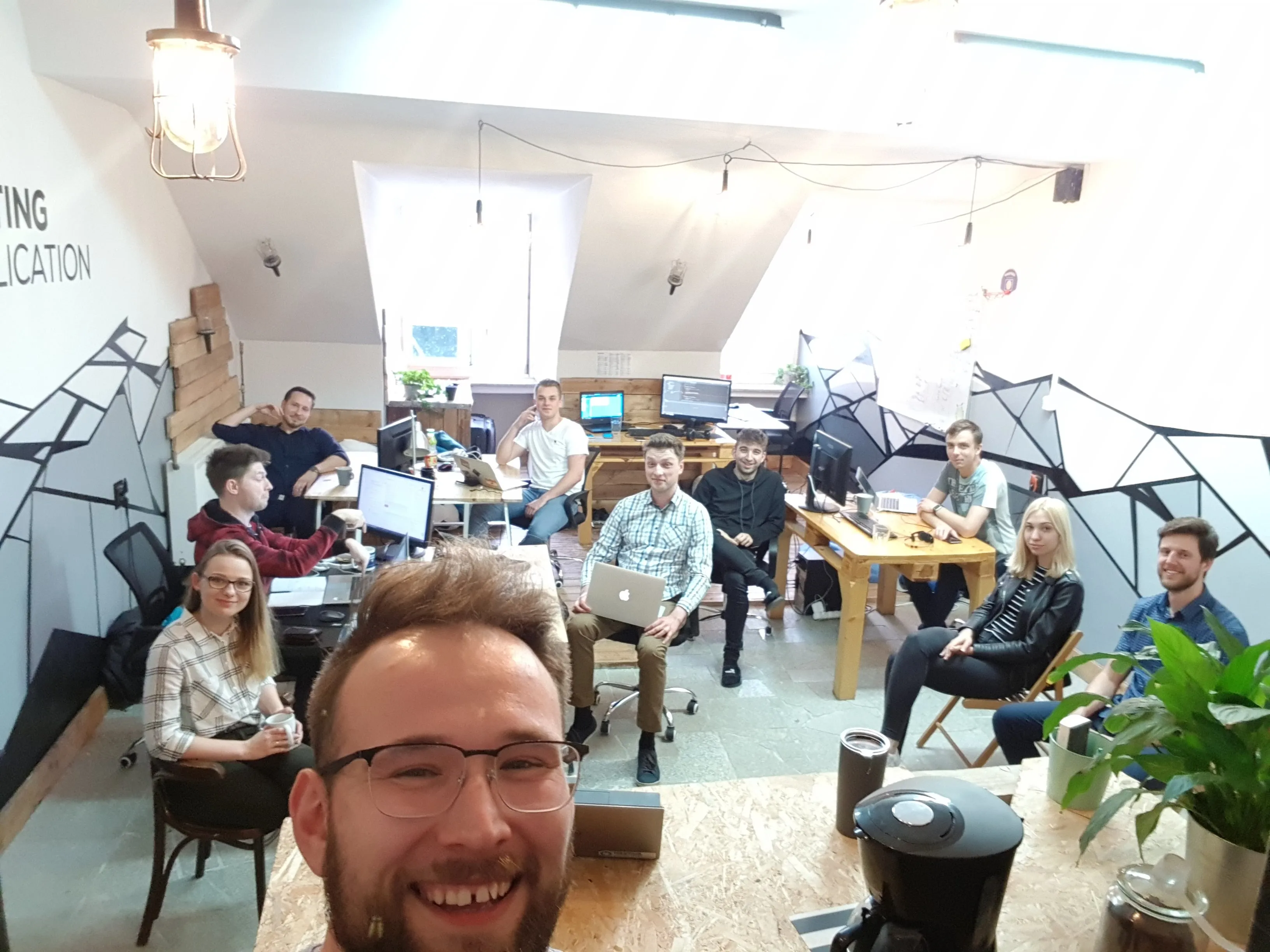 A group of ten people in an office, some working at computers, and one man with glasses takes a smiling selfie.