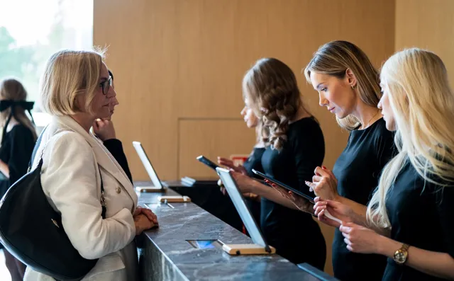 Kobieta w białym płaszczu rozmawiająca z trzema recepcjonistkami w czarnych ubraniach przy lady recepcyjnej.