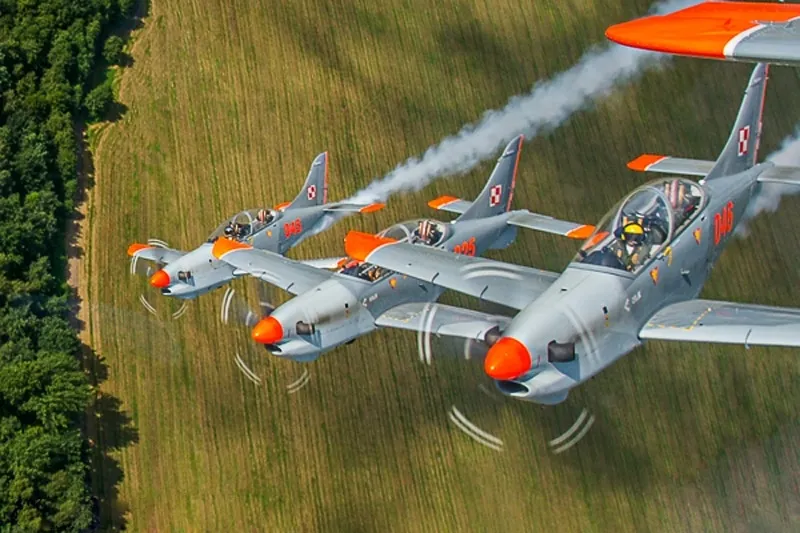 Three gray military planes with orange noses and wings flying in formation over a field next to a forest.