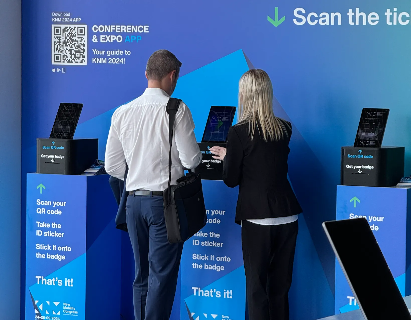 Dwóch osób rejestrujących się na Kongresie Nowej Mobilności 2024 przy stanowiskach z czytnikami kodów QR w celu pobrania identyfikatora.