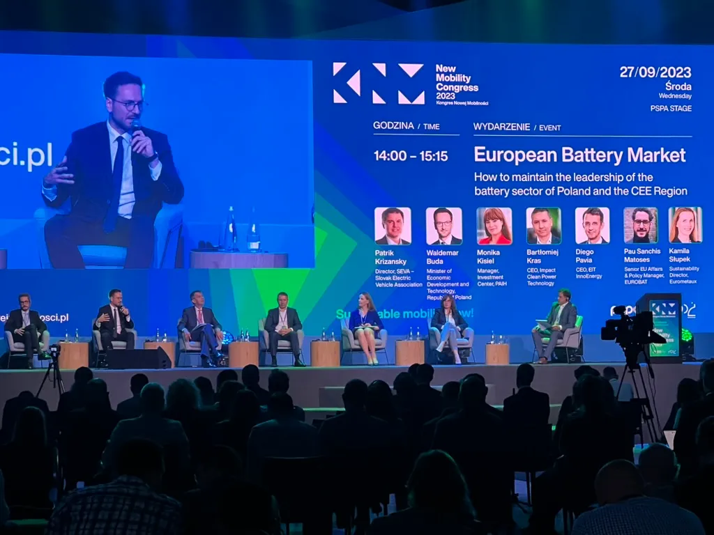 Discussion panel during New Mobility Congress 2023 about the European battery market, with seven speakers on stage and a large screen presenting event details.