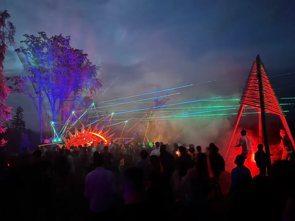 Crowd of people at night at outdoor light show with lasers and red and blue illuminations on trees and structures.