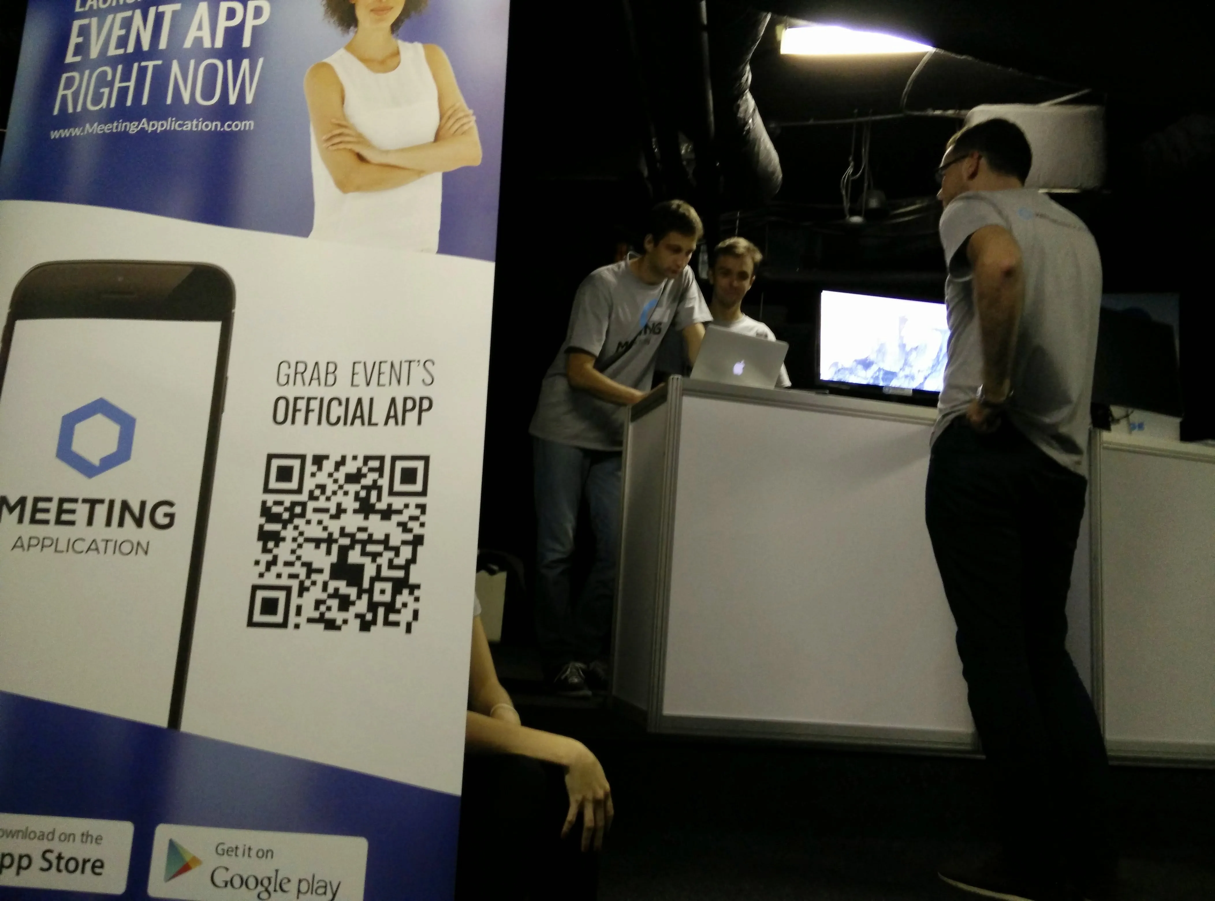 Meeting Application promotional stand with a large banner and three men standing by the stand with a laptop.