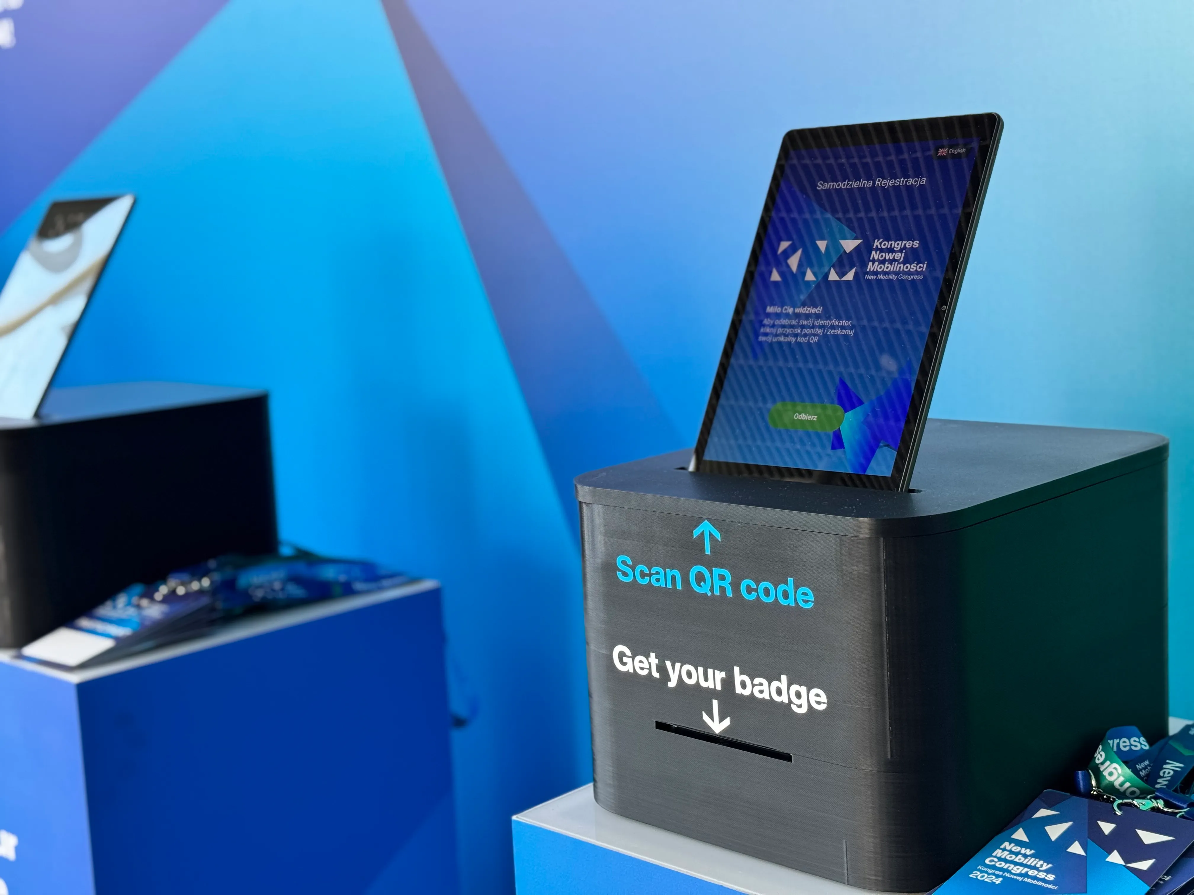 Stojak na identyfikatory z tabelą do skanowania kodu QR i tabletem wyświetlającym ekran Kongres Nowej Mobilności.