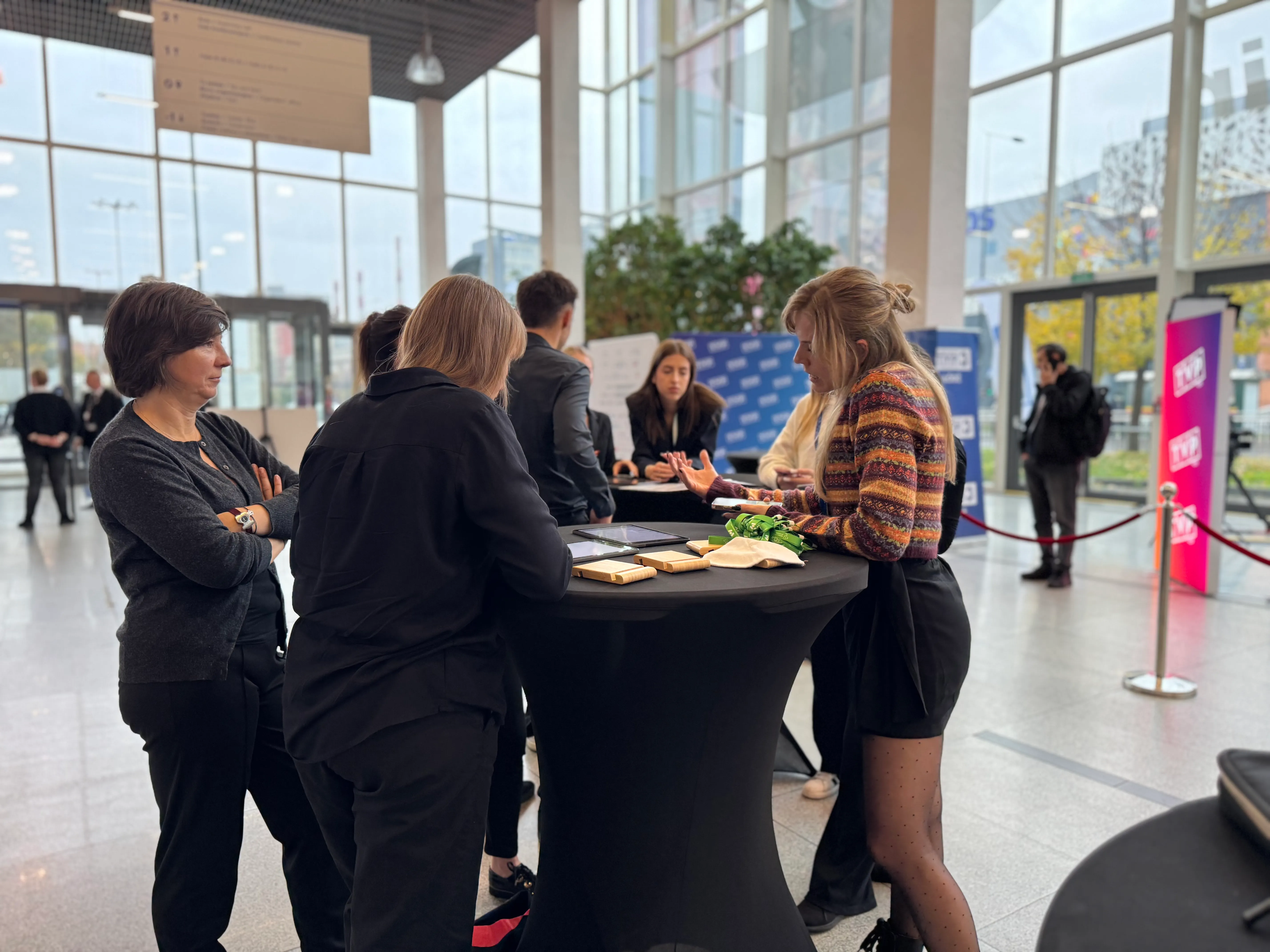 Grupa ludzi rozmawiająca wokół wysokiego okrągłego stołu w jasnym holu z dużymi oknami.