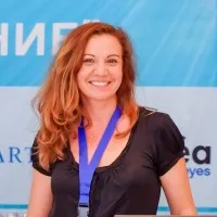 Uśmiechnięta kobieta z rudymi falującymi włosami nosząca niebieską wstążkę na szyi i czarną bluzkę.