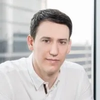 Mężczyzna z krótkimi ciemnymi włosami noszący białą koszulę, siedzący w jasnym pomieszczeniu z oknami w tle.