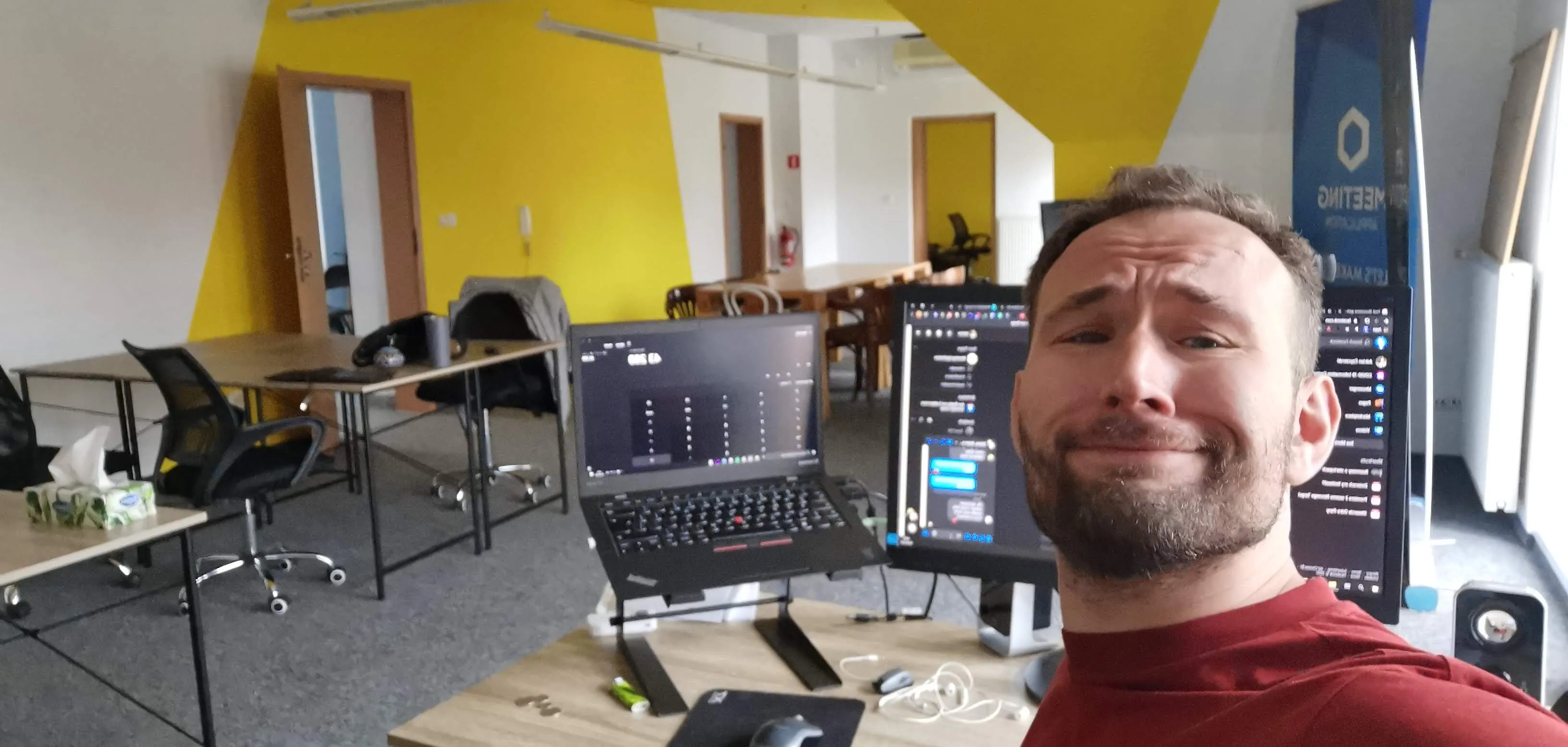 A man taking a selfie at a desk with two monitors and a laptop in a modern office with yellow walls.