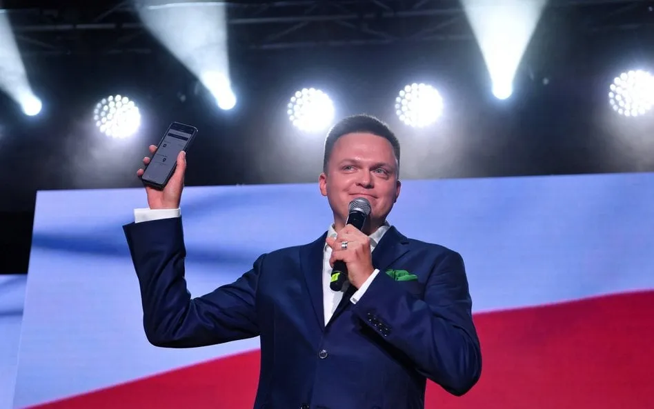 Man in navy blazer holding microphone and raising smartphone on stage with bright lighting and flag in background.