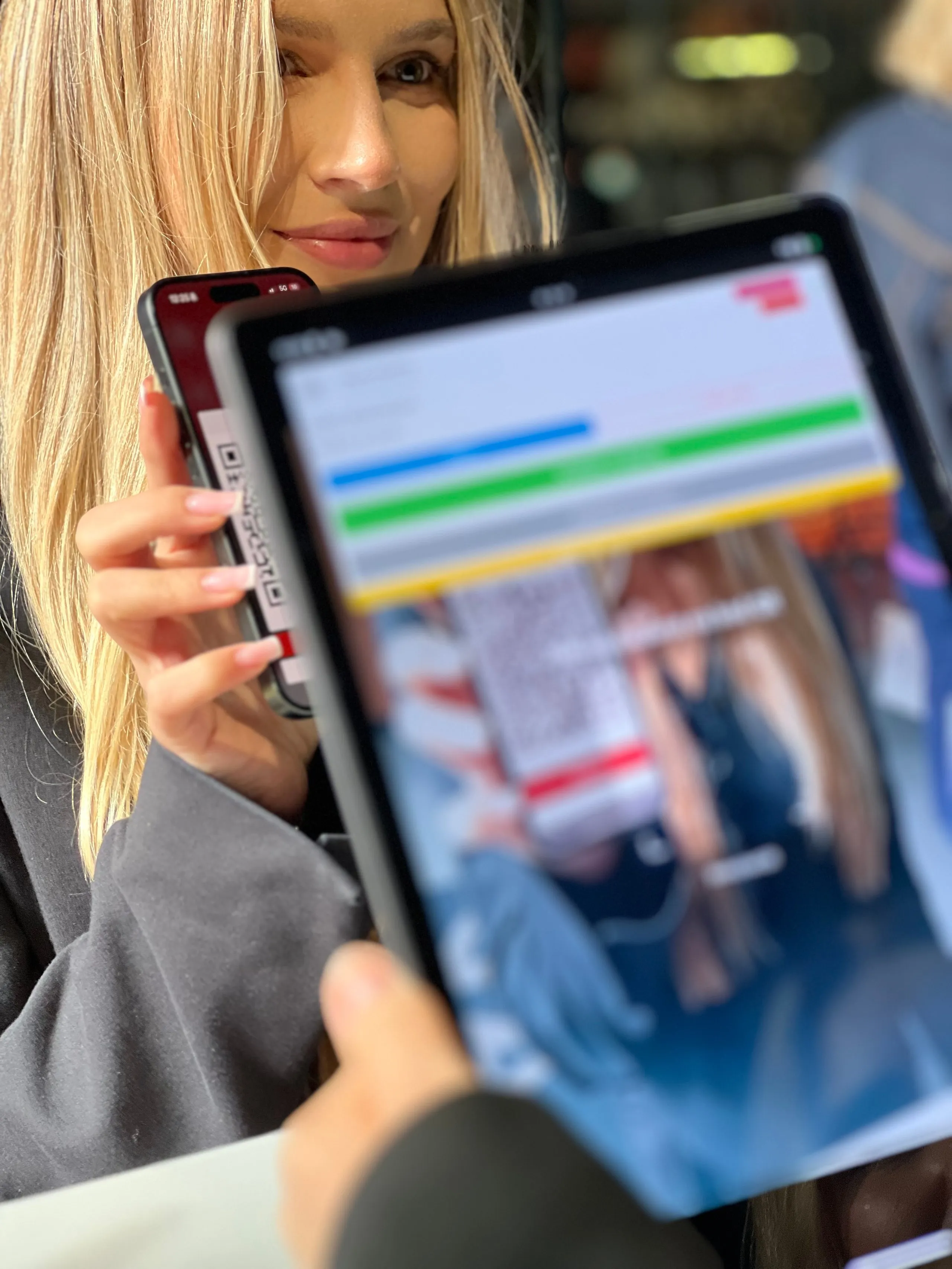 Young woman showing smartphone screen with QR code while someone else scans it using a tablet.