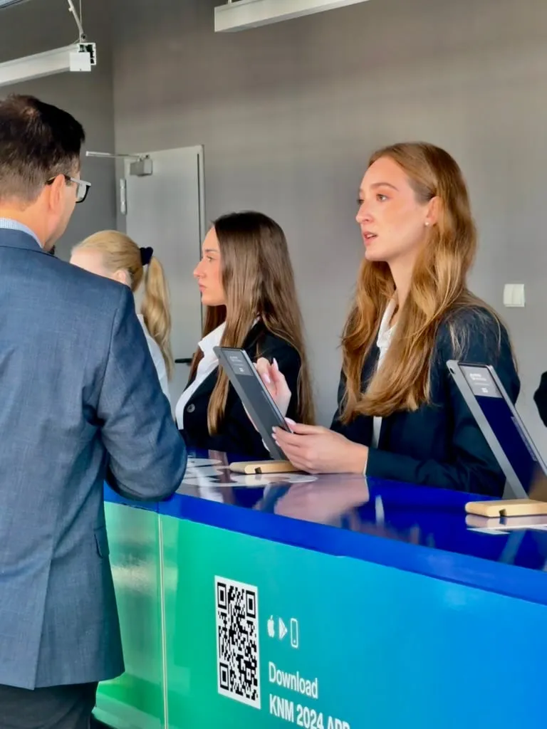 Two women behind counter serving a man, holding tablets at blue-green booth with QR code and text 'Download KNM 2024 APP'.