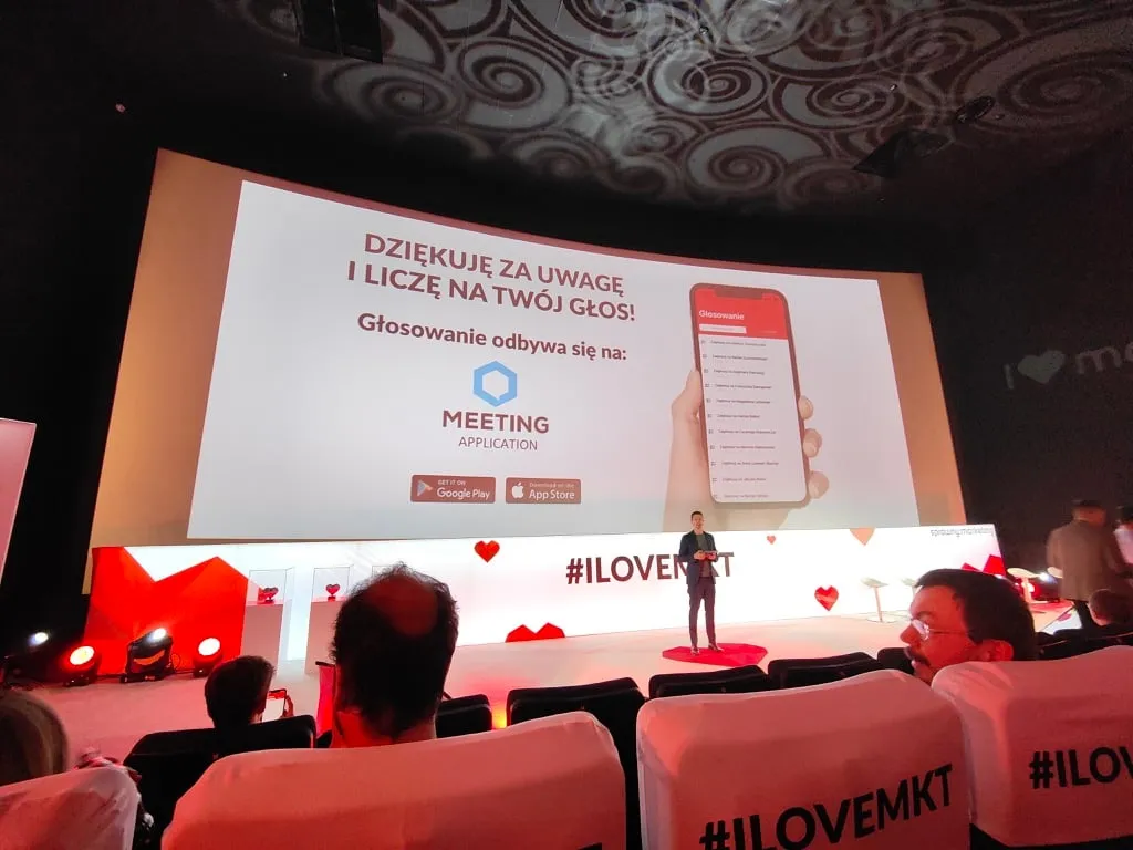 Speaker on stage in conference hall presenting voting via Meeting app, screen visible with instructions and Google Play and App Store icons.