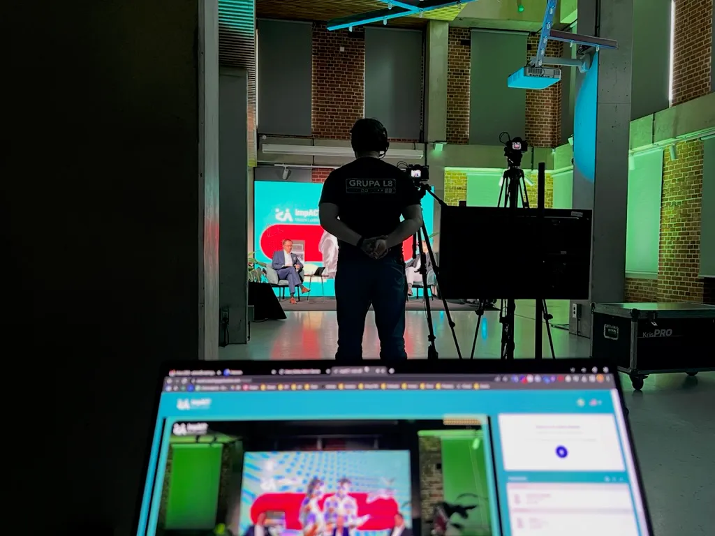 Person with camera and tripod standing in recording studio, laptop in foreground showing live broadcast.