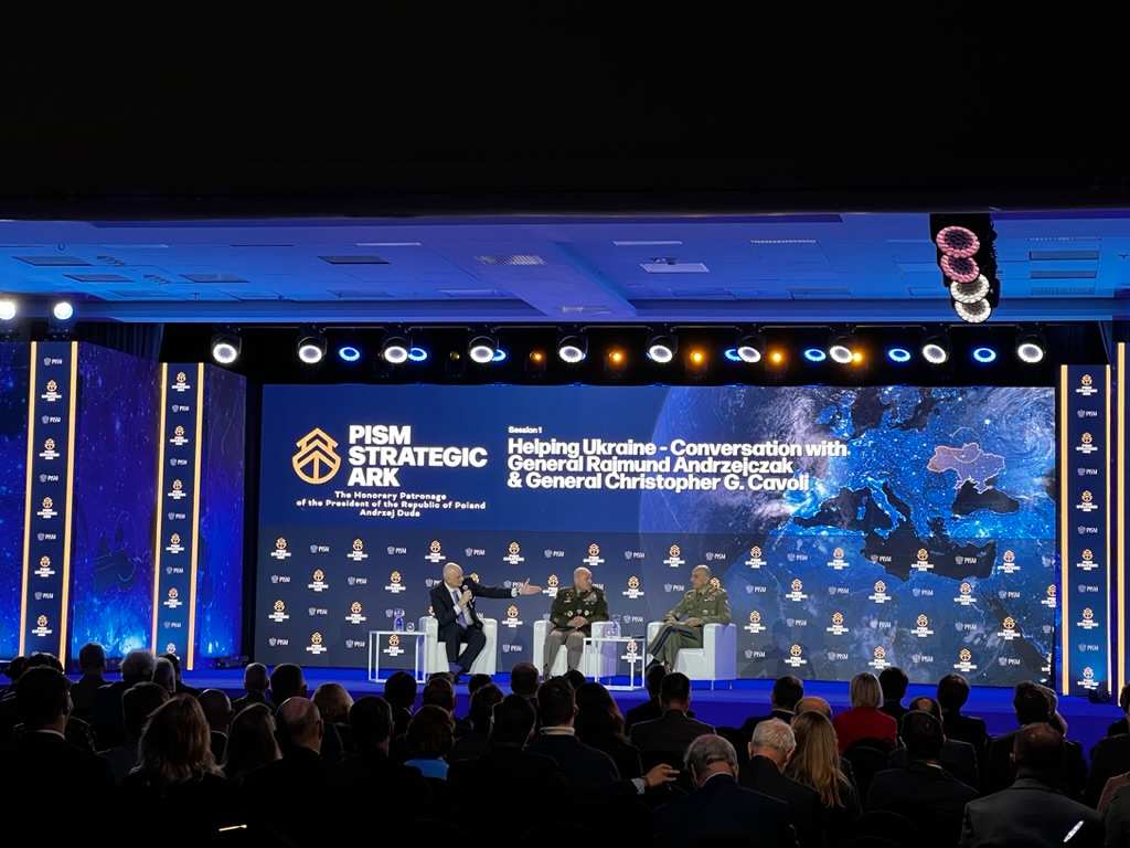 Panel discussion with three men on stage at PISM Strategic Ark conference, with map of Europe and text supporting Ukraine in background.