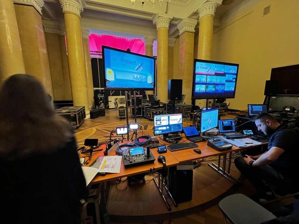 Person sitting at table with multiple monitors and technical equipment in hall with columns and large screen displaying presentation about Poland.