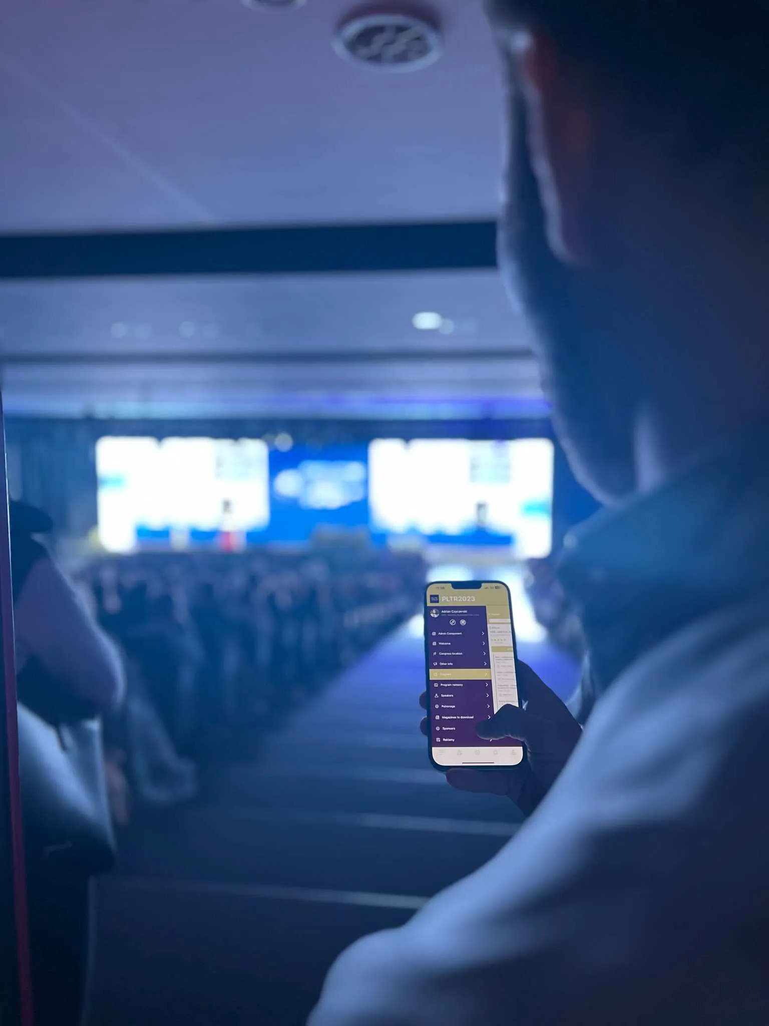 Person holding smartphone displaying conference app during event in hall with auditorium.