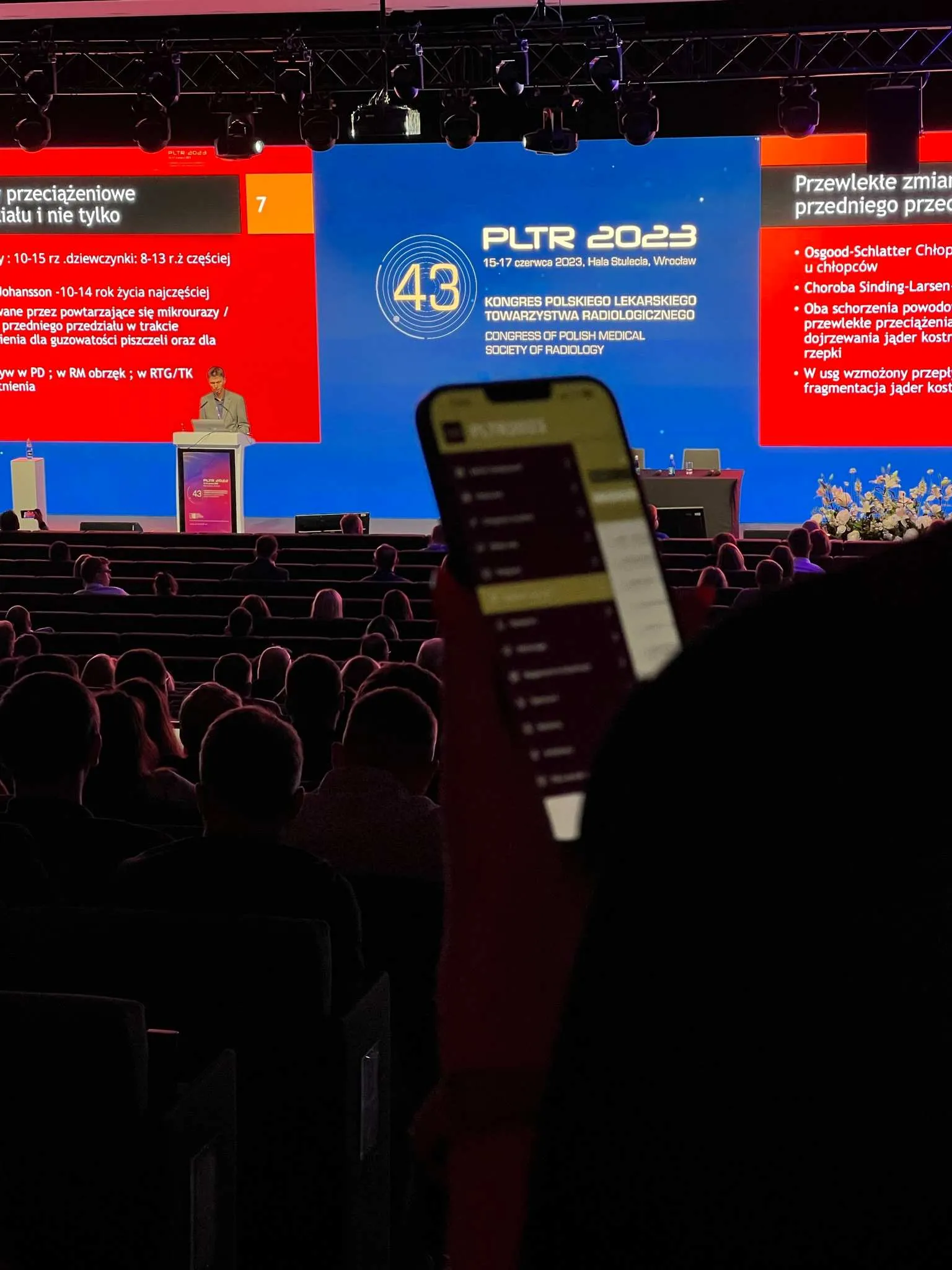 Speaker speaking in front of large screen with PLTR 2023 logo during Polish radiological society congress, participant viewing conference program on phone.