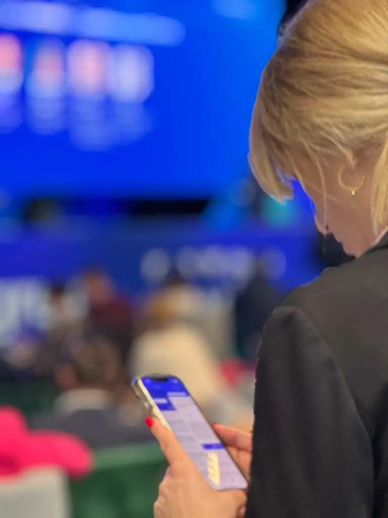 Blonde woman holding smartphone showing sent chat messages, sitting against blurred audience and large blue screen background.