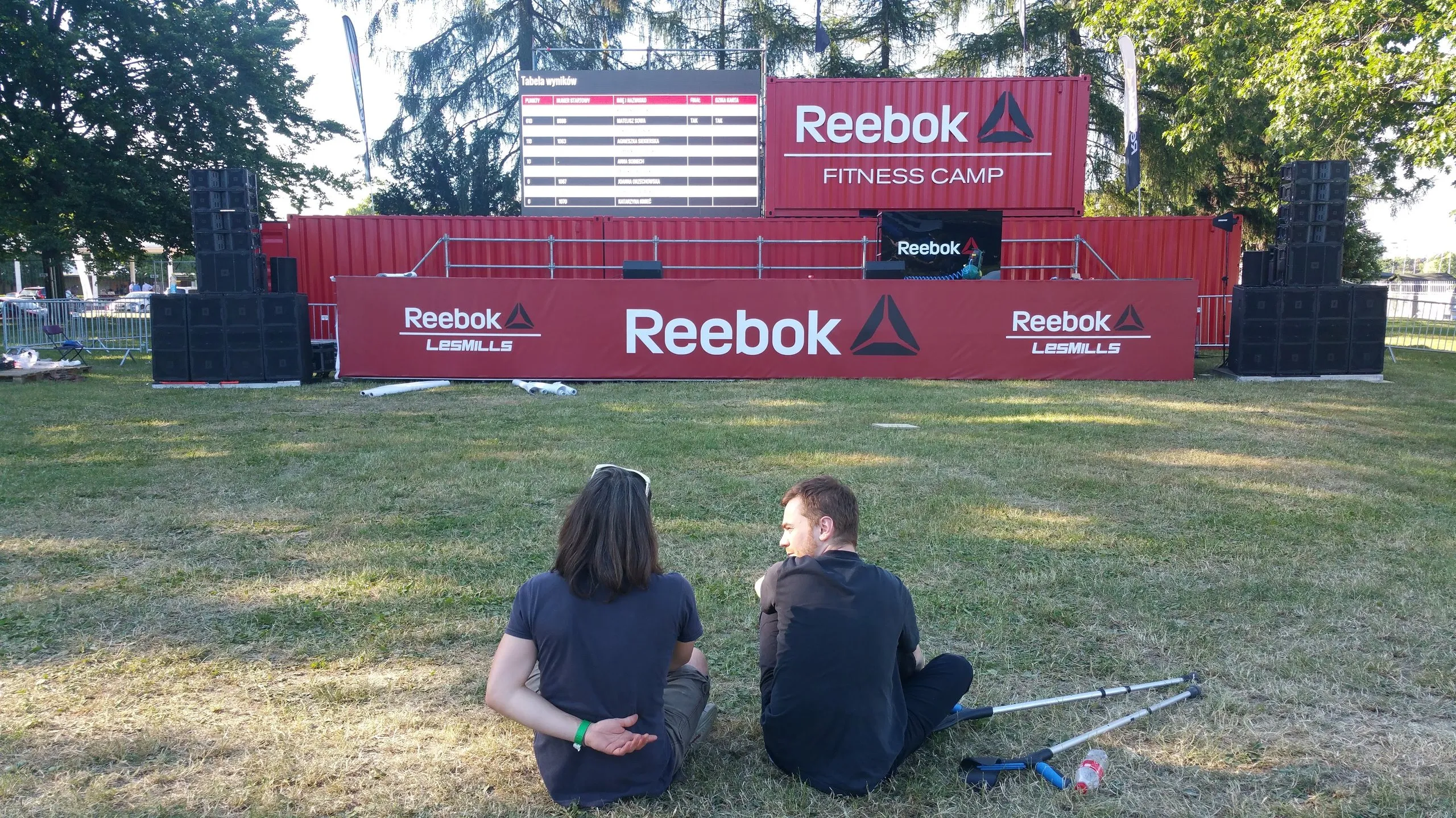 Two people sitting on grass in front of stage with red Reebok advertising banners at outdoor festival.