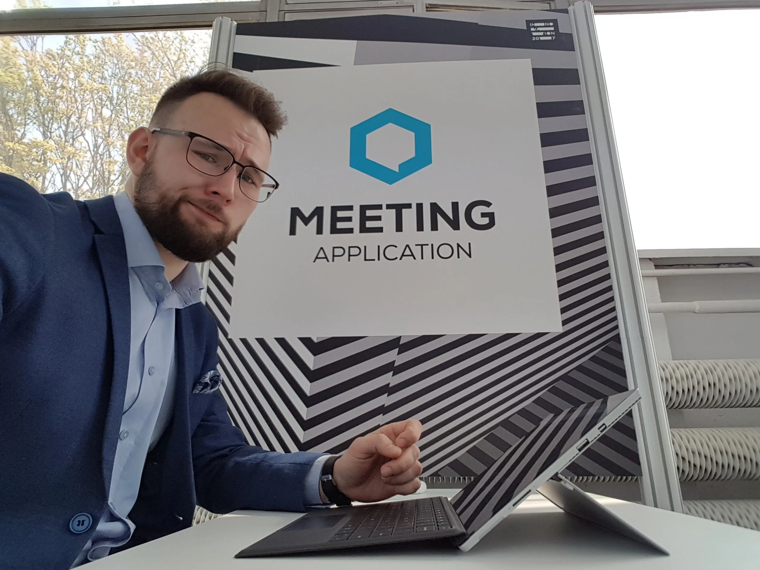 Man in glasses and navy blazer working on laptop, sitting at table in front of board with text Meeting Application.