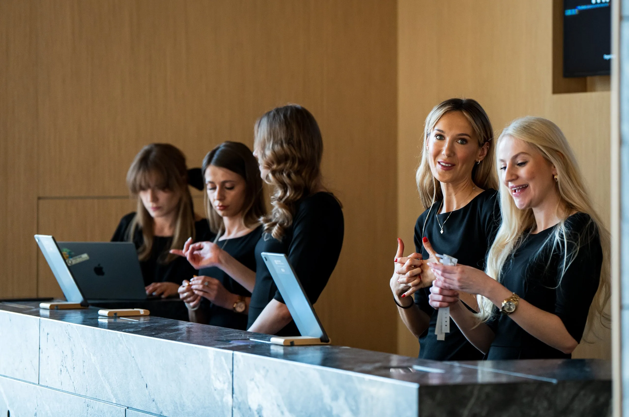 Five women behind reception desk with two laptops and tablets, two smiling during conversation.