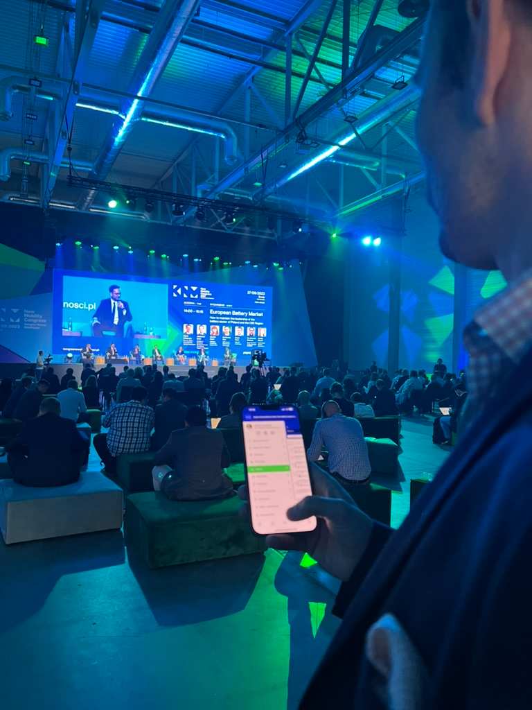 Man holding phone showing a list, during conference with audience and panel discussion in background.