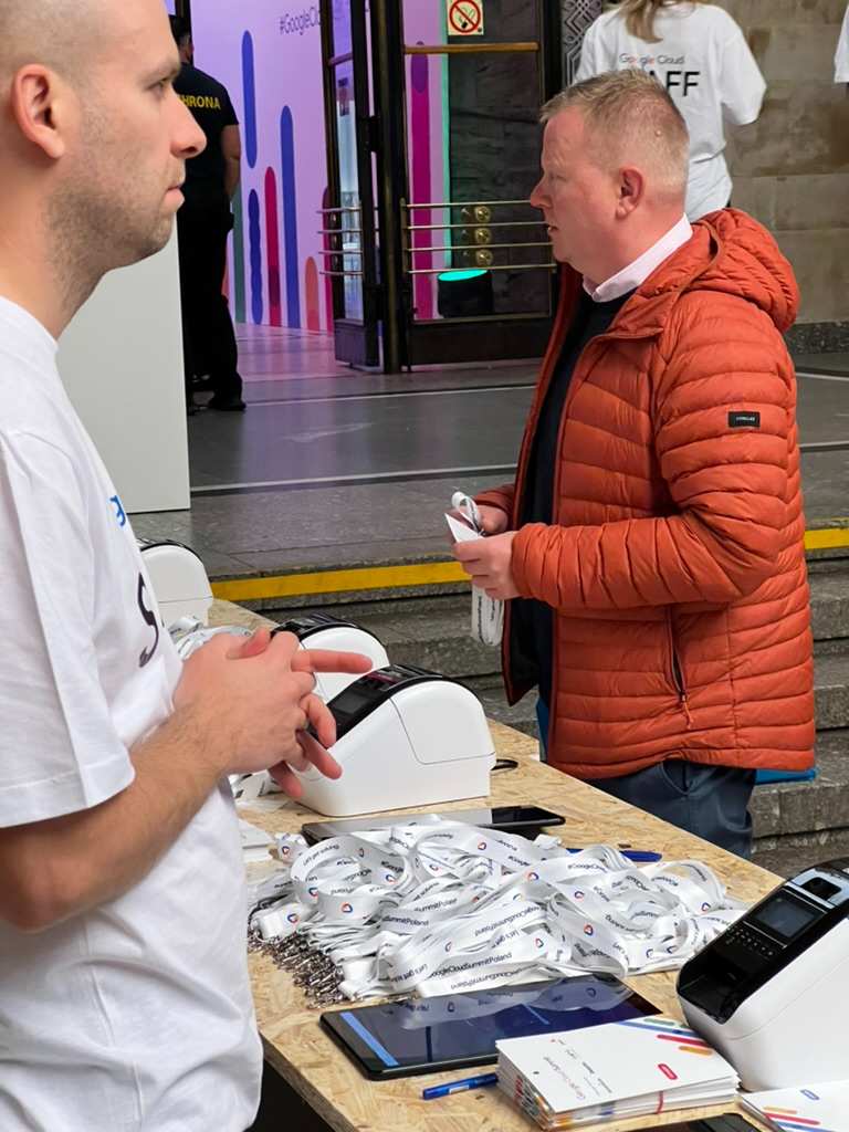 Man in white shirt standing at table with ID badges and printers, second man in orange jacket holding ID badge.