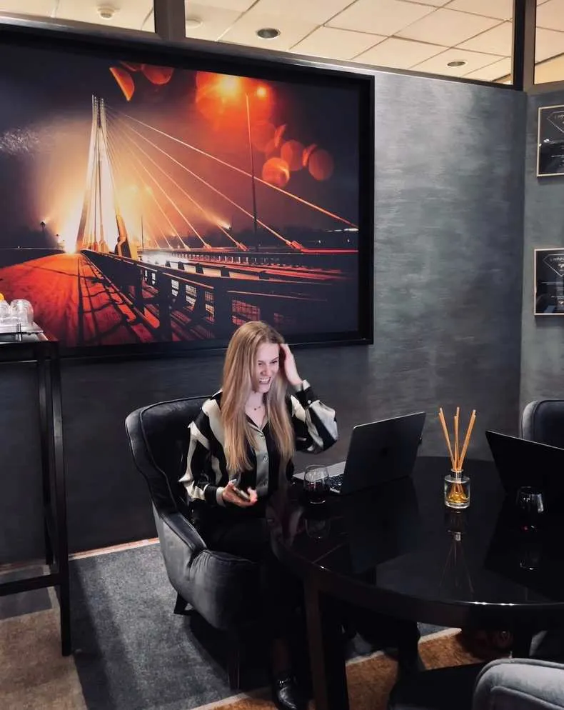 Woman sitting at black table with laptop, glass of red wine nearby, against large photo of bridge at night.