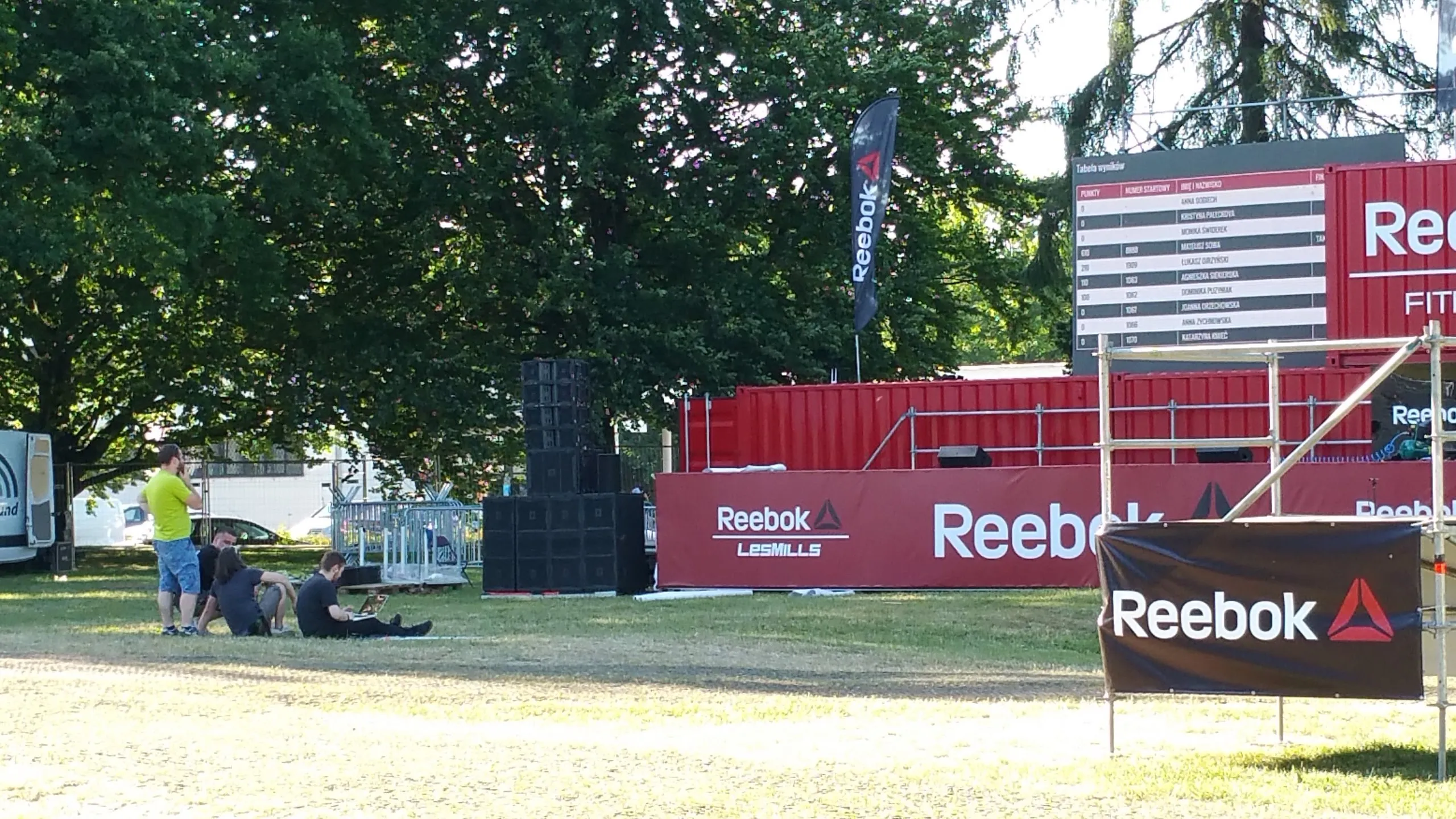 Group of people sitting and standing on grass next to a stage and banners with the Reebok logo at an outdoor event.