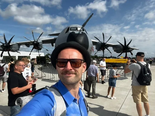 Man in glasses and a hat taking a selfie against the background of a military aircraft with four propellers and a group of people at an air show.