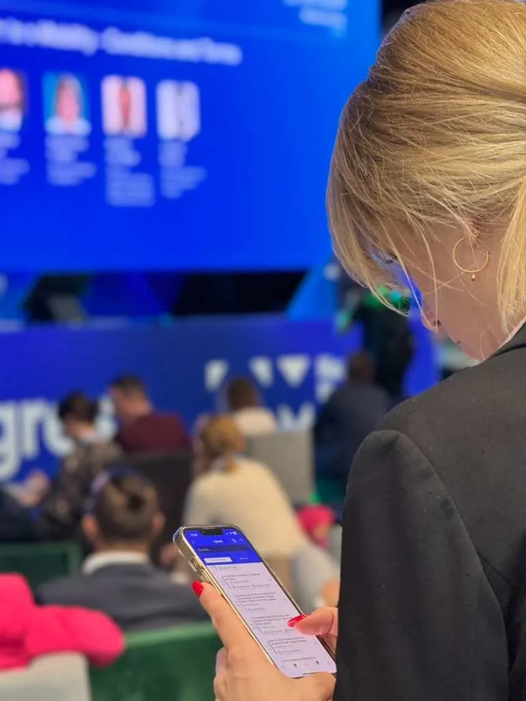 A woman with light hair and red nail polish uses a smartphone during an event with people sitting in front of a blue screen.