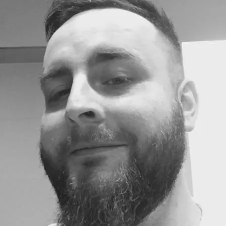 Zbliżenie twarzy mężczyzny z krótkimi włosami i gęstą brodą, uśmiechającego się delikatnie.